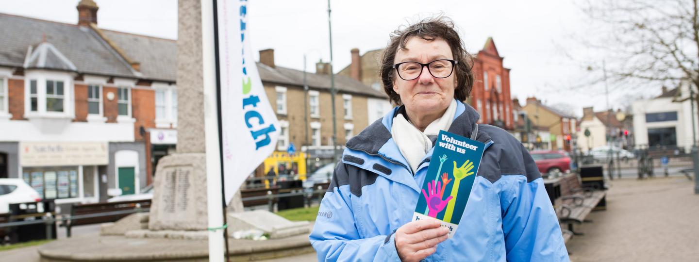 Healthwatch volunteer holding a leaflet on volunteering and stood in front of a Healthwatch banner